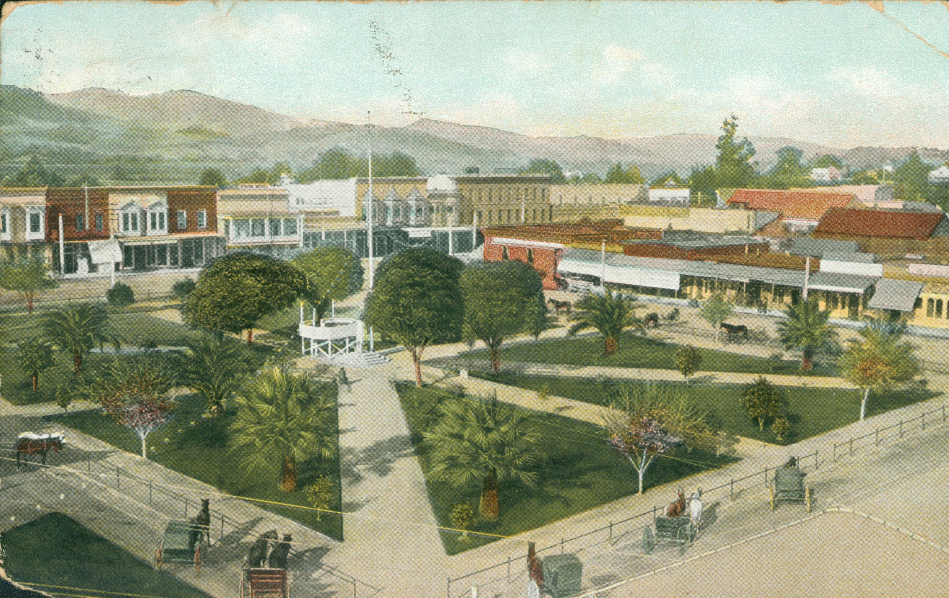 Shows a park planted with several trees and surrounded by carriages and buildings